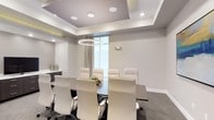 This professional conference room presents a refined and contemporary design suited for formal meetings and executive gatherings. The space is anchored by a substantial dark boardroom table that comfortably accommodates multiple attendees, surrounded by high-back white leather chairs that convey a sense of sophistication and comfort. Overhead, a striking coffered ceiling adds architectural depth and dimension, complemented by recessed lighting and a statement circular pendant light that serves as the room's focal point. The neutral color palette — featuring soft whites, warm grays, and dark accents — creates a calm, focused atmosphere conducive to productivity. A large window dressed with floor-length sheer drapes allows natural light to filter into the space, enhancing its bright and airy feel. A built-in credenza along one wall provides functional storage and surface space, while subtle wall-mounted fixtures contribute to the layered lighting design. Dark-toned carpeting grounds the room and provides a polished contrast to the lighter furnishings. Overall, this conference room balances elegance with functionality, making it an ideal setting for professional presentations, board meetings, and collaborative discussions.