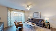 Located at 833 S University Blvd in Mobile, Alabama, this single-floor residence offers a thoughtfully designed layout with a cohesive, modern aesthetic throughout. The living room serves as the heart of the space, featuring wood-look flooring, a ceiling fan, and large sliding glass doors framed by decorative teal-patterned curtains that open to a private balcony, allowing natural light to pour in and providing outdoor views. The galley-style kitchen is efficiently arranged with white cabinetry, light quartz-style countertops, wood-look tile flooring, and black appliances including a range and built-in microwave, maximizing functionality within a streamlined footprint. The unit includes two well-sized bedrooms, each appointed with plush carpeting, ceiling fans, and windows dressed with patterned curtains. Both bedrooms offer a comfortable, restful ambiance with ample space for full bedroom furnishings. Two full bathrooms complement the bedrooms, each featuring wood-look flooring, white vanity cabinetry with quartz-style countertops, and shower-tub combinations. The overall design palette of neutral tones accented with teal and navy details creates a cohesive, inviting atmosphere throughout the entire residence.