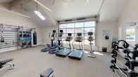 Located in Solana Beach, California, this dedicated fitness facility at 309 Solana Hills Dr is a comprehensively outfitted gym designed to support a wide range of workout routines. The space is defined by its striking vaulted ceiling with exposed beams, multiple skylights, and a large floor-to-ceiling glass garage-style door that floods the interior with natural light and provides open views of the surrounding neighborhood.

The cardio zone features a lineup of treadmills, elliptical machines, and a stationary bike, each equipped with individual display monitors. Adjacent to this area, a dedicated strength training section houses a multi-station cable machine, a Smith machine, and a full rack of dumbbells arranged on tiered stands, complemented by adjustable benches to accommodate a variety of exercises.

A functional training wall lines one end of the room, outfitted with a modular storage system holding medicine balls, kettlebells, resistance tools, and a heavy punching bag — ideal for dynamic and circuit-style training. Full-length mirrors along one wall enhance the sense of space and allow for form monitoring during workouts.

The interior finishes are clean and minimal, with neutral-toned carpet flooring, white walls, and exposed HVAC ductwork running along the ceiling. A ceiling fan provides additional air circulation. The overall environment is bright, well-ventilated, and purpose-built for serious fitness use.