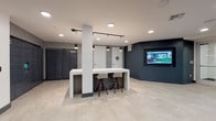 Located at 1935 Stockton Blvd in Sacramento, California, this contemporary single-floor space serves as a welcoming lobby and package management hub. The interior is defined by a clean, modern aesthetic that blends light and dark tones throughout.

The lounge area is bright and inviting, featuring floor-to-ceiling glass walls that flood the space with natural light and offer views of the surrounding exterior. Comfortable high-back lounge chairs are arranged around a warm wood coffee table atop a textured area rug, creating an approachable and relaxed seating environment.

Adjacent to the lounge, the package and mail area is outfitted with a full bank of dark smart parcel lockers, providing secure and convenient package retrieval. A sleek white high-top counter with bar-height seating sits centrally in the space, ideal for quick tasks or casual use, and is illuminated by pendant lighting suspended from an overhead track system.

A wall-mounted flat-screen display is recessed into a bold dark accent wall, adding a tech-forward touch to the space. Light-toned tile flooring extends throughout, enhancing the sense of openness, while recessed ceiling lighting ensures consistent illumination across all areas. Glass-paneled double doors connect the lobby to additional interior spaces, maintaining visual continuity and an open, airy feel throughout.