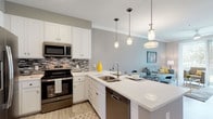 This thoughtfully designed single-floor residence offers a cohesive blend of modern finishes and functional living spaces. The kitchen is a standout feature, showcasing crisp white shaker cabinetry, a mosaic tile backsplash, gleaming quartz countertops, and a full suite of stainless steel appliances including a French door refrigerator, electric range with over-the-range microwave, and dishwasher. A peninsula with bar seating and pendant lighting above creates a natural transition into the open-concept dining and living area, where light wood-look tile flooring and crown molding add a refined touch. The living room is bright and inviting, with large sliding glass doors that frame serene waterfront views and allow natural light to fill the space. The bedroom is generously proportioned, featuring plush carpeting, recessed lighting, and ample room for a king-size bed and additional furnishings. The bathroom is finished with large-format tile surround in the tub and shower combination, complemented by a decorative mosaic accent strip and a quartz vanity. A dedicated in-unit laundry area with a full-size washer and dryer provides added convenience, completing this well-rounded and move-in-ready living space.