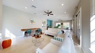 This single-story clubhouse or community room showcases a spacious, open-plan layout designed for both socializing and relaxation. Light-toned hardwood flooring runs throughout, complementing the crisp white walls and lending the interior a clean, airy feel. Ceiling fans are positioned at intervals across the space, ensuring comfortable airflow throughout.

A central bar-height island with a white stone countertop serves as the social hub of the space, surrounded by vibrant teal and green upholstered barstools with wood frames. A bold teal accent wall frames the beverage station nearby, which includes a coffee and drink service area with a sink — a practical amenity for gatherings. White cabinetry below the island adds ample storage.

Adjacent to the bar area, a generous lounge zone features white sofas and armchairs arranged around a pair of round wood accent tables. A floor-to-ceiling tile-clad wall houses a sleek, wide linear fireplace, flanked by large windows that draw in natural light and offer views of the surrounding greenery.

A built-in wooden desk along one wall provides a functional workspace with teal chairs. French doors open toward an outdoor area, further connecting the interior to the exterior environment. The overall ambiance is coastal, fresh, and inviting.