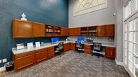 Located at 5720 Forest Park Rd in Dallas, Texas, this single-floor business center is designed to support productivity in a polished, well-appointed environment. The space makes a striking first impression with its dramatically high ceilings and oversized arched transom windows that flood the interior with natural light, creating an open and airy atmosphere despite its compact footprint.

The workstation area is thoughtfully outfitted with wrap-around, warm-toned wood cabinetry and built-in shelving that lines the walls, providing generous storage and display space for books and reference materials. Multiple computer workstations with ergonomic chairs are arranged along the continuous countertop surface, accompanied by a multifunction printer, making the area fully equipped for day-to-day business needs. Wi-Fi access is also available in this area.

A dedicated conference area offers a sleek, dark rectangular table surrounded by modern ergonomic chairs, ideal for small meetings or collaborative work sessions. Large French doors with floor-to-ceiling glass panels and transom windows above allow views to the exterior and invite additional natural light into the meeting space. The interiors are finished with a refined combination of teal accent walls, neutral grey tones, and patterned carpeting, lending the space a professional yet welcoming character.