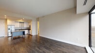 This single-floor residence showcases a thoughtfully designed open-concept layout, where the living area flows seamlessly into the kitchen. Dark hardwood flooring extends throughout the main living spaces, creating a warm and cohesive aesthetic against neutral beige walls and crisp white trim.

The kitchen is a standout feature, outfitted with white shaker-style cabinetry, including glass-front upper cabinets, dark granite countertops, and a light-toned subway tile backsplash. A central island with a speckled stone surface provides additional prep space and casual seating. Stainless steel appliances, including a French door refrigerator with ice maker, dishwasher, microwave, and range, complete the modern setup.

The bedrooms continue the dark hardwood flooring, with one bedroom featuring a large floor-to-ceiling window offering expansive city views and a ceiling fan with integrated lighting. A walk-in closet and en-suite bathroom are also present, with the primary bath featuring a glass-enclosed walk-in shower, dark granite vanity, and light tile flooring. A second bathroom includes a soaking tub with subway tile surround and a backlit vanity mirror, adding a refined touch throughout.