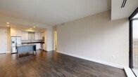 This single-floor residence showcases a thoughtfully designed open-concept layout, where the living area flows seamlessly into the kitchen. Dark hardwood flooring extends throughout the main living spaces, creating a warm and cohesive aesthetic against neutral beige walls and crisp white trim.

The kitchen is a standout feature, outfitted with white shaker-style cabinetry, including glass-front upper cabinets, dark granite countertops, and a light-toned subway tile backsplash. A central island with a speckled stone surface provides additional prep space and casual seating. Stainless steel appliances, including a French door refrigerator with ice maker, dishwasher, microwave, and range, complete the modern setup.

The bedrooms continue the dark hardwood flooring, with one bedroom featuring a large floor-to-ceiling window offering expansive city views and a ceiling fan with integrated lighting. A walk-in closet and en-suite bathroom are also present, with the primary bath featuring a glass-enclosed walk-in shower, dark granite vanity, and light tile flooring. A second bathroom includes a soaking tub with subway tile surround and a backlit vanity mirror, adding a refined touch throughout.