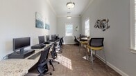 Located at 1589 Starlight Cv in Tarpon Springs, Florida, this single-floor business center is thoughtfully designed to support both focused work and casual collaboration. The space is finished with soft gray walls, crisp white crown molding, and patterned carpet flooring, creating a clean and professional atmosphere throughout.

The main work area is lined with granite-topped desks accommodating multiple computer workstations, each equipped with monitors, keyboards, and ergonomic task chairs. This setup provides ample workspace for productivity and is well-suited for a range of computing needs. Pendant and flush-mount lighting fixtures complement the natural light that streams in through multiple windows dressed with white blinds, keeping the environment bright and inviting.

A dedicated lounge corner offers a high-top table with bar-height seating in accented yellow chairs, providing a more informal setting for collaboration or casual use. A cozy accent chair and decorative teal side table add warmth to an adjacent sitting nook, enhancing the overall comfort of the space.

The walls are adorned with curated artwork, including abstract aquatic paintings and a sculptural metal wall piece, contributing to a polished yet welcoming aesthetic. French glass-paned doors connect the areas, adding architectural character while maintaining an open, airy feel throughout the space.