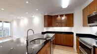 This single-floor residence offers a thoughtfully designed layout encompassing one bedroom and one bathroom. The kitchen is a standout feature, showcasing warm medium-toned wood cabinetry that extends from floor to ceiling, providing generous storage throughout. Upper cabinets include glass-front display doors with integrated lighting, adding a refined touch to the space. Dark granite countertops provide a striking contrast against the cabinetry and white walls, lending the kitchen a polished, contemporary aesthetic. A stainless steel undermount sink is paired with a modern pull-down faucet, combining functionality with style. The kitchen also includes a stainless steel dishwasher, seamlessly integrated into the cabinetry. Light hardwood flooring runs throughout, brightening the space and complementing the overall design. Recessed lighting and a flush ceiling fixture ensure the kitchen is well-lit, while crown molding along the ceiling adds an elegant architectural detail. The clean white walls and efficient layout create a bright, functional environment suited for everyday living.
