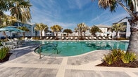 This outdoor amenity space centers around a generously sized freeform swimming pool with clear turquoise water, designed to evoke a coastal resort atmosphere. The pool's curved, organic shape adds visual interest and is complemented by gently sloping entry points. Surrounding the pool is a broad, well-finished paver deck constructed from large-format stone tiles in neutral gray and beige tones, providing ample space for relaxation and circulation.

A row of lounge chairs and tilt umbrellas in soft aqua tones lines the perimeter, offering shaded seating throughout the day. Mature palm trees are thoughtfully positioned across the deck and surrounding landscape beds, reinforcing a tropical aesthetic while providing natural shade. Low ornamental plantings, including agave and ornamental grasses, frame the edges of the space.

Adjacent to the pool, a single-story clubhouse structure features coastal-style board-and-batten siding in crisp white and beige tones, large multi-pane windows, and a covered outdoor porch with ceiling fans, extending the usable space for gathering. The entire pool area is enclosed by a black metal fence, ensuring security while maintaining open sightlines to the surrounding residential buildings.