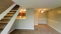 This two-story residence presents a practical and open layout designed for comfortable everyday living. The main level showcases a seamlessly connected living and dining area, finished with wall-to-wall carpeting that adds warmth underfoot. Neutral gray-toned walls provide a versatile backdrop throughout the space, complemented by clean white baseboards and trim that frame each room with a crisp, finished look.

A defining architectural feature of the main level is the kitchen pass-through, which opens directly onto the living area, promoting an open, connected feel between the cooking and gathering spaces. The kitchen, partially visible from the living area, features warm wood-toned cabinetry and a light countertop surface. A staircase runs along one wall, leading to the upper floor and adding vertical dimension to the space.

The living area is equipped with a ceiling fan, supporting air circulation throughout the room. Overhead lighting fixtures provide consistent illumination across the main level. The layout makes efficient use of the available footprint, with each area flowing naturally into the next, making the overall space feel open and well-organized.
