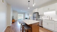 This thoughtfully designed 1-bedroom, 1-bathroom residence offers a functional and modern open-concept layout across a single floor. The kitchen serves as a centerpiece of the living space, boasting crisp white shaker cabinetry, a white subway tile backsplash, quartz countertops, and a full suite of stainless steel appliances including a refrigerator, dishwasher, electric range, and microwave. A wood-toned kitchen island with a quartz top and seating for two is illuminated by stylish pendant lighting, making it ideal for casual dining or meal prep. Warm hardwood-style flooring flows throughout the main living areas, adding cohesion and warmth to the space. The living room is bright and airy, featuring a large sliding glass door with vertical blinds that allows abundant natural light and provides outdoor views. A dedicated closet offers convenient storage. The bedroom is a calm, carpeted retreat with a large window framing outdoor greenery, providing a quiet and comfortable environment. A full bathroom features a white vanity with quartz countertop, a tub-shower combination, and hardwood-style flooring. A bold teal interior door adds a distinctive architectural accent throughout the home.