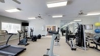 Located at 150 Horizon Trl in Garner, NC, this dedicated fitness center offers a well-appointed space designed to support a comprehensive workout routine. The single-floor facility is bright and welcoming, with recessed panel lighting and a ceiling fan that ensure a comfortable environment during exercise sessions. The walls are finished in a clean, light gray tone that contributes to an open and airy atmosphere. A large wall mirror spans one side of the room, visually expanding the space and allowing users to monitor their form during workouts. The facility is equipped with a range of cardio machines, including elliptical trainers and additional aerobic equipment, alongside a multi-station strength-training machine and weight benches. The neutral-toned flooring is well-suited for the demands of a fitness environment. A wall-mounted television adds an entertainment option for those looking to stay engaged during longer cardio sessions. French doors with glass panes provide a welcoming entryway, adding a touch of architectural charm to this functional space. Overall, the fitness center at 150 Horizon Trl presents a clean, organized, and fully equipped environment suitable for a wide range of fitness activities.