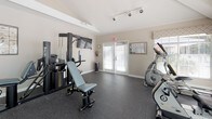 This single-level fitness center offers a thoughtfully arranged workout space designed to support a full range of exercise activities. The room features durable rubber flooring throughout, providing a safe and functional surface for equipment use and free movement. Track lighting mounted on the ceiling ensures consistent, even illumination across the space.

Natural light is a standout element, with multiple large windows dressed in patterned Roman shades and horizontal blinds lining one wall and offering views of an outdoor pool area. A large floor-to-ceiling mirror is installed along another wall, visually expanding the space and supporting form-checking during workouts.

The fitness center is equipped with a Vectra multi-station strength training system, an adjustable weight bench, treadmills, a stationary upright bike, a recumbent bike, and an elliptical machine. A wall-mounted flat-screen television is positioned near the strength equipment for entertainment during workouts. French doors with frosted panel windows provide access to the exterior, contributing additional natural light. A fire extinguisher and posted instructional guides complete the practical, safety-conscious setup of this well-maintained fitness facility.