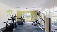 This single-level fitness center is thoughtfully designed to accommodate a well-rounded exercise routine. The main workout area is bright and welcoming, characterized by large arched windows that flood the space with natural light and offer views of surrounding greenery, creating an energizing atmosphere. A bold yellow-green accent wall adds a vibrant focal point to the room.

The cardio zone is well-stocked with multiple True-brand treadmills and elliptical trainers, as well as stationary bikes, providing ample options for aerobic exercise. The strength training section features a multi-station cable machine, a bench press, and additional resistance equipment suited for a full-body workout. A wall-mounted flat-screen television is positioned for convenient viewing during exercise sessions.

Underfoot, a multi-toned teal and blue modular carpet tiles add visual interest while providing cushioning throughout the workout space. Nature-themed artwork decorates the walls, contributing to the facility's fresh and motivating ambiance. A textured white ceiling with recessed ventilation completes the clean aesthetic.

Adjacent to the main gym floor, a separate glass-enclosed lounge area with light tile flooring and seating offers a comfortable space to rest or gather before or after workouts, accessible through French-style glass-panel doors.