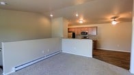 This single-floor space showcases an open-concept design that seamlessly connects the living area to the kitchen. The living area is finished with plush wall-to-wall carpeting and features a baseboard heating system along the perimeter, providing efficient climate control. Recessed lighting in the ceiling offers clean, functional illumination throughout the space.

A half-wall partition separates the living area from the kitchen, creating a natural division while maintaining an open, airy feel. The kitchen is well-appointed with medium-toned wood cabinetry, granite-style countertops, and a full suite of stainless steel appliances, including a refrigerator, microwave, and range. The kitchen features durable hardwood-style flooring, which contrasts with the carpeted living space and adds a practical, easy-to-maintain surface in the cooking area.

The neutral, warm beige tones on the walls complement the overall color palette of the space, creating a cohesive and welcoming atmosphere. The straightforward, functional layout makes efficient use of the available space, offering a comfortable and practical living environment suited for everyday use.