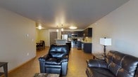 This single-floor space is designed with an open-concept layout that seamlessly connects the living room, dining area, and kitchen into one cohesive environment. The living room anchors the space with plush black leather recliners and dark stone-topped side tables, creating a comfortable and functional seating area. Hardwood-style flooring runs throughout, lending warmth and visual continuity across all zones.

The kitchen is well-appointed with dark cabinetry that extends to the upper walls, offering generous storage. Stainless steel appliances, including a gas range, microwave, and refrigerator, provide a modern, practical edge. A kitchen island with a light countertop adds prep space and serves as a natural divider between the kitchen and living areas. The sink is positioned along the far wall beneath upper cabinetry, complemented by a tile backsplash.

Adjacent to the kitchen, a compact dining area accommodates a dark wood table and chairs, ideal for everyday meals. Pendant lighting above the island and flush-mount ceiling fixtures provide layered illumination throughout. The neutral yellow-toned walls and consistent flooring unify the space, making it feel open and well-organized.