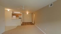 This single-floor space offers a practical and functional open-concept layout that seamlessly connects the main living area with the kitchen. The living room features warm, neutral-toned walls and is finished with plush carpeting, creating a comfortable and inviting atmosphere. A ceiling fan centered overhead provides both lighting and airflow to the space.

The kitchen is situated adjacent to the living area and is accessible via a breakfast bar-style counter, which also serves as a natural divider between the two spaces. The kitchen is outfitted with warm wood-toned cabinetry, a white refrigerator, and a range with overhead ventilation. The kitchen floor transitions to tile, providing a practical and easy-to-clean surface.

Durable hardwood-style flooring extends through a portion of the main living area, adding visual contrast to the carpeted sections. Recessed lighting and a utility light fixture supplement the ceiling fan light, ensuring the space is well-lit throughout.

A hallway visible from the main living area leads to additional rooms, suggesting a thoughtful and organized floor plan. The clean white trim and neutral wall tones throughout give the space a cohesive, move-in-ready appearance. Overall, the layout balances open living with defined functional zones.