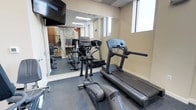 This fitness center is a thoughtfully appointed exercise space designed to meet essential workout needs in a clean, functional environment. The room is outfitted with quality cardio equipment, including a treadmill and an elliptical trainer by Life Fitness, providing users with reliable options for cardiovascular exercise. A wall-mounted flat-screen television is positioned for easy viewing during workouts, enhancing the overall experience. Full-length mirrors line one wall, visually expanding the space while allowing users to monitor their form. The flooring features durable, rubberized gym-grade material that provides both safety and comfort underfoot. Natural light filters in through a window, contributing to a bright and energizing atmosphere alongside the recessed ceiling lighting. The neutral-toned walls and drop ceiling lend the space a clean, professional aesthetic. The mirrored wall reflects additional equipment visible in the background, suggesting further workout options are available within the same area. Overall, this fitness center is a practical and well-maintained space suited for daily exercise routines.
