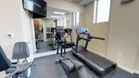 This fitness center is a thoughtfully appointed exercise space designed to meet essential workout needs in a clean, functional environment. The room is outfitted with quality cardio equipment, including a treadmill and an elliptical trainer by Life Fitness, providing users with reliable options for cardiovascular exercise. A wall-mounted flat-screen television is positioned for easy viewing during workouts, enhancing the overall experience. Full-length mirrors line one wall, visually expanding the space while allowing users to monitor their form. The flooring features durable, rubberized gym-grade material that provides both safety and comfort underfoot. Natural light filters in through a window, contributing to a bright and energizing atmosphere alongside the recessed ceiling lighting. The neutral-toned walls and drop ceiling lend the space a clean, professional aesthetic. The mirrored wall reflects additional equipment visible in the background, suggesting further workout options are available within the same area. Overall, this fitness center is a practical and well-maintained space suited for daily exercise routines.