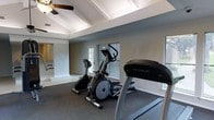 This fitness room offers a thoughtfully designed workout environment within a single-story layout. The space is characterized by its vaulted ceiling with elegant coffered detailing and crown molding, lending a sense of architectural refinement uncommon in typical gym settings. A ceiling fan is centrally mounted overhead, providing air circulation during workouts.

The room is furnished with quality cardio equipment, including a TRUE-brand treadmill, an elliptical trainer, and a stationary upright bike, all positioned on durable carpeted flooring. The neutral gray wall tones create a calm, focused atmosphere conducive to exercise.

Natural light filters in through multiple windows along the walls, as well as through a glass-paneled door that leads to an exterior area, keeping the space bright and open. A partial view into an adjacent room is visible through an arched opening, suggesting a connected layout. The combination of refined architectural details and functional fitness equipment makes this a distinctive and comfortable space for daily exercise.