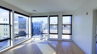 This single-floor space offers a clean, contemporary aesthetic defined by an abundance of natural light and open sightlines. The main living area is anchored by a striking bank of large, floor-to-ceiling windows framed in bold black trim, spanning nearly the entire exterior wall. These expansive windows flood the interior with sunlight throughout the day, casting warm light across the smooth, light-toned hardwood flooring. The corner window placement further enhances the sense of openness, offering panoramic views of the surrounding urban streetscape, including neighboring residential buildings and city rooftops. The walls are finished in crisp white, complementing the minimalist design and allowing the natural light to reflect evenly throughout the space. The ceiling is smooth and unadorned, with a standard HVAC vent integrated discreetly overhead, maintaining the clean lines of the interior. The hardwood floors run consistently across the space, adding warmth and continuity to the overall aesthetic. Together, the modern window design, neutral finishes, and generous light create an inviting, airy atmosphere well-suited to a variety of uses.