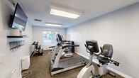 Located at 703 Broadridge Ln in Saint Peters, Missouri, this fitness center offers a focused and fully equipped exercise environment within a single, efficiently arranged room. The space features neutral light-gray walls, textured carpet flooring, and bright flush-mounted panel lighting that ensures a well-lit atmosphere throughout the day. Multiple windows with slatted blinds allow natural light to filter in while providing views of an outdoor pool and surrounding amenity area, adding an open, airy feel to the room.

The exercise room is outfitted with a selection of True-brand commercial-grade equipment, including a treadmill, a recumbent stationary bike, and a multi-function cable strength machine. A dedicated free weights area features a rack of dumbbells and an adjustable weight bench positioned near a wall-mounted mirror, supporting strength training and flexibility exercises. Along one wall, built-in shelving and hooks provide convenient storage for personal items, fitness accessories, and supplies. A wall-mounted flat-screen television adds an entertainment option during workouts. The room is climate-controlled with visible HVAC vents and includes a waste bin for easy upkeep, ensuring a clean and comfortable environment for all users.
