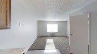 This single-floor studio offers a straightforward, open layout designed for efficient use of space. The main living area features a combination of hard flooring near the entrance and wall-to-wall carpeting throughout the central and rear portions of the room, providing a practical transition between zones. Neutral grey walls create a clean, understated backdrop that allows for flexible furnishing and décor choices.

A centrally positioned window along the far wall admits natural light, casting brightness across the room and contributing to an airy feel. Beneath the window sits a wall-mounted HVAC unit, supplemented by baseboard heating that runs along the perimeter walls, ensuring year-round climate control. The textured ceiling adds subtle visual interest to the otherwise minimalist space.

A doorway to the right offers access to an additional area, likely a bathroom or closet, rounding out the functional layout. The overall ambiance is simple and neutral, presenting a blank canvas well-suited for personalization. The space is practical and manageable, ideal for a single occupant seeking a no-frills, low-maintenance living environment.