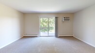 Located at 1440 S Neltnor Blvd in West Chicago, IL, this 2-bedroom, 1-bathroom unit offers a functional single-floor layout with a comfortable and practical design throughout. The living room serves as the central gathering space, featuring plush carpet, baseboard heating, a wall-mounted air conditioning unit, and a large sliding glass door that opens to a balcony overlooking a well-maintained lawn with mature trees — bringing in generous natural light and pleasant outdoor views. The kitchen is positioned adjacent to the living area, equipped with a gas range and wood-toned cabinetry, providing an efficient workspace. A hallway connects the main living areas to the two bedrooms, each carpeted and outfitted with a ceiling fan and light fixture. The primary bedroom is notably spacious, offering a double-door closet and a window with views of the greenery outside. The second bedroom also benefits from natural light via its own window. The full bathroom includes a tub and shower combination with a white surround. Neutral wall tones and clean white trim carry throughout the unit, creating a cohesive and move-in ready atmosphere. The property's grounds feature open green spaces, walkways, and tree coverage, enhancing the overall setting.