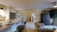 This single-floor residence is designed with an open-concept layout that seamlessly connects the living and dining areas, creating a cohesive and functional living space. The living area features plush wall-to-wall carpeting in a warm neutral tone, contributing to a comfortable and inviting ambiance. A cushioned sofa anchors the space, complemented by a dark wood display cabinet that adds a touch of classic elegance.

The dining area is highlighted by a peninsula-style breakfast bar with bar stool seating, ideal for casual dining or entertaining. A modern pendant light hangs above the bar, providing focused task lighting and a stylish focal point. The adjacent wall features a bold black-and-white framed artwork, adding visual interest to the otherwise soft, neutral walls.

A hallway visible from the main living area leads to additional rooms, including what appears to be a bedroom, offering a sense of depth and privacy within the home. The consistent use of light-toned walls and carpeting throughout creates a bright, airy atmosphere, while the thoughtful mix of furnishings and decor lends the space a well-appointed, move-in ready feel.