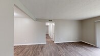 This single-floor unit offers a clean, modern aesthetic highlighted by warm wood-look luxury vinyl plank flooring that runs throughout every room. The open living area is bathed in natural light from a large sliding glass door that opens to a view of the surrounding greenery, creating a bright and welcoming atmosphere. A textured popcorn ceiling and neutral warm-gray walls provide a consistent, cohesive backdrop across all spaces.

The galley-style kitchen is thoughtfully designed with crisp white shaker cabinets, light stone-patterned countertops, a stainless steel refrigerator and range, a built-in dishwasher, and a double-basin sink — offering ample storage and prep space in an efficient layout. A decorative tile backsplash adds a subtle design accent.

A central hallway connects the living area to the bedrooms, each of which features the same wood-look flooring, neutral walls, and a window providing natural light and outdoor views. The bathroom is straightforward and functional, equipped with a shower-tub combination, a single vanity with a framed mirror, and brushed nickel fixtures. The overall layout is practical and well-organized, making excellent use of the available space throughout the unit.