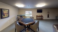Located at 3594 N Cederblom St in Coeur d'Alene, Idaho, this single-floor community amenity space is designed for recreation and social gathering. The main game room is generously sized, with neutral carpet flooring, neutral-toned walls, and flush-mounted lighting that keeps the space bright and welcoming. At the center of the room sits a full-size pool table with a green felt surface, complemented by a wall-mounted cue rack nearby. Along one side, a shuffleboard table is paired with a high-top bar table and stool, while framed automotive-themed artwork decorates the adjacent wall, adding a stylish, casual character to the room. A dedicated card and game area features an octagonal poker table surrounded by upholstered chairs, a wall-mounted flat-screen television, and a small console table for accessories. A separate seating area includes a round high-top table with bar-height chairs and a compact mini-refrigerator, providing a convenient refreshment station. A pass-through window offers a connection to an adjacent area. The space also includes a private meeting room furnished with a rectangular table and leather-style chairs, suitable for small group gatherings or quiet conversations. Together, these areas create a versatile, well-appointed community hub ideal for both leisure and casual meetings.
