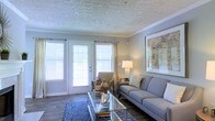 This single-story space showcases a thoughtfully designed living room that balances comfort and style. The room is anchored by a set of French doors flanked by large windows fitted with white horizontal blinds and flowing drapery panels, allowing generous natural light to fill the interior while offering views to the outdoors.

The ceiling features a distinctive textured pattern with crown molding trim, adding architectural character to the space. Dark hardwood-style flooring grounds the room and provides a rich contrast to the lighter wall tones and furnishings.

The living area is arranged around a glass-topped coffee table set atop a patterned blue area rug, complemented by a grey upholstered sofa dressed with accent pillows. A cushioned armchair and side table with a table lamp create a secondary seating nook near the French doors. A large framed art piece adorns one wall, contributing to the polished, curated aesthetic of the room.

Overall, the space presents a clean, neutral palette with warm accents, creating an inviting and functional living environment that is both practical and visually appealing.