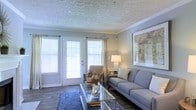 This single-story space showcases a thoughtfully designed living room that balances comfort and style. The room is anchored by a set of French doors flanked by large windows fitted with white horizontal blinds and flowing drapery panels, allowing generous natural light to fill the interior while offering views to the outdoors.

The ceiling features a distinctive textured pattern with crown molding trim, adding architectural character to the space. Dark hardwood-style flooring grounds the room and provides a rich contrast to the lighter wall tones and furnishings.

The living area is arranged around a glass-topped coffee table set atop a patterned blue area rug, complemented by a grey upholstered sofa dressed with accent pillows. A cushioned armchair and side table with a table lamp create a secondary seating nook near the French doors. A large framed art piece adorns one wall, contributing to the polished, curated aesthetic of the room.

Overall, the space presents a clean, neutral palette with warm accents, creating an inviting and functional living environment that is both practical and visually appealing.