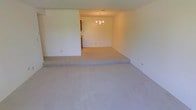 This single-floor space showcases a distinctive split-level layout, where a raised carpeted area steps down into a sunken living space — a classic architectural feature that adds depth and character to the interior. Both levels are finished with plush, neutral-toned wall-to-wall carpeting that runs seamlessly throughout, contributing to a warm and comfortable ambiance. The sunken area is anchored by a decorative chandelier overhead, lending an elegant focal point to the space. The walls are finished in a clean, neutral tone, providing a versatile backdrop suitable for a variety of design styles. A partial wall divides the main living area from an adjacent space, hinting at a connected kitchen or dining area visible in the background. The overall layout feels open and airy, with the stepped transition between levels offering a natural sense of separation between functional zones without sacrificing the open-plan feel. The space is well-suited for both relaxed everyday living and entertaining, with the sunken design creating an intimate gathering area within the broader floor plan.