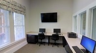 This single-floor space serves as a dedicated business center or computer lab, thoughtfully designed to support productivity and ease of use. The room features an L-shaped built-in workstation with a light-colored countertop and dark cabinetry below, offering ample desk surface for multiple users simultaneously. Two computer monitors with keyboards and ergonomic rolling office chairs are positioned side by side, providing comfortable individual workstations. A large flat-screen television is mounted prominently on the wall above the desk, ideal for presentations or shared viewing. A printer is conveniently stationed at the end of the counter, completing the functional workspace setup. The room benefits from natural light streaming in through multiple windows dressed with neutral blinds and a decorative patterned valance, lending a bright and welcoming atmosphere. The walls are finished in a clean, neutral tone, and the floor is covered with a neutral-colored carpet, contributing to a quiet and focused environment. The generous ceiling height adds a sense of openness to the space, making it feel comfortable despite its compact footprint. Overall, this business center is a practical, well-appointed space suited for work, research, or collaborative tasks.