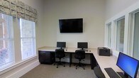 This single-floor space serves as a dedicated business center or computer lab, thoughtfully designed to support productivity and ease of use. The room features an L-shaped built-in workstation with a light-colored countertop and dark cabinetry below, offering ample desk surface for multiple users simultaneously. Two computer monitors with keyboards and ergonomic rolling office chairs are positioned side by side, providing comfortable individual workstations. A large flat-screen television is mounted prominently on the wall above the desk, ideal for presentations or shared viewing. A printer is conveniently stationed at the end of the counter, completing the functional workspace setup. The room benefits from natural light streaming in through multiple windows dressed with neutral blinds and a decorative patterned valance, lending a bright and welcoming atmosphere. The walls are finished in a clean, neutral tone, and the floor is covered with a neutral-colored carpet, contributing to a quiet and focused environment. The generous ceiling height adds a sense of openness to the space, making it feel comfortable despite its compact footprint. Overall, this business center is a practical, well-appointed space suited for work, research, or collaborative tasks.