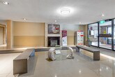 This building lobby offers a functional and welcoming entrance experience across a generously sized single-floor layout. The space is defined by gleaming large-format white ceramic tile flooring, accented by strategically placed carpet runners at key transition points, including the main entrance and elevator bay.

Upon entry through the automatic glass sliding doors, residents and visitors are greeted by a bright foyer with recessed and flush-mount ceiling lighting. A mailroom station is conveniently positioned near the entrance for easy access. The seating area features a mix of upholstered sofas and patterned accent chairs arranged around a decorative fireplace mantle, providing a comfortable communal gathering space.

A striking stacked-stone accent wall adds warmth and texture to the lounge area, complemented by framed artwork displayed nearby. The elevator core is finished with elegant stone-clad wall panels surrounding two stainless steel elevator doors, lending a polished, modern aesthetic. Additional building amenity signage and an intercom system are integrated throughout.

The neutral warm-toned walls, combined with consistent overhead lighting, create a clean and inviting ambiance throughout the lobby. The overall design balances practicality with comfort, presenting a professional and well-maintained common area for building residents.