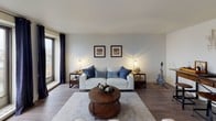 This single-floor residence is thoughtfully designed with a cohesive navy and neutral color palette throughout. The living area is a welcoming space anchored by light upholstered seating, hardwood-style flooring, and large windows dressed in navy grommet curtains that frame city views and invite generous natural light. Framed artwork and warm accent lighting contribute to a comfortable, contemporary ambiance.

The kitchen stands out with its bold chalkboard-painted cabinetry, creating a striking contrast against the light countertops and stainless steel appliances, which include a refrigerator, range, microwave, and dishwasher. A decorative area rug adds a playful touch underfoot.

The bedroom offers a restful retreat with large windows overlooking the cityscape, a ceiling fan, and ample closet storage with bifold doors. A dresser with a mirror, wall-mounted television, and bedside lighting complete the well-appointed space.

The bathroom features tile flooring, a dark-finish vanity with under-sink storage, an over-toilet cabinet for additional storage, and a shower-tub combination. The overall design of the unit balances practicality with a modern, urban aesthetic.