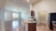 This well-appointed single-floor residence offers a thoughtfully designed open-concept layout that seamlessly connects the kitchen, dining, and living areas. The kitchen serves as a functional centerpiece, featuring rich dark-wood cabinetry, granite countertops, a breakfast bar peninsula with an integrated sink, and a full suite of stainless steel appliances including a refrigerator, range, microwave, and dishwasher. Track lighting runs overhead, complementing pendant fixtures above the bar for focused task illumination. The expansive living area is bathed in natural light from multiple windows and is finished with gleaming hardwood-style flooring that flows throughout the main spaces. Soaring ceilings and exposed utility piping lend an industrial-loft character to the space, while a ceiling fan adds comfort. Two full bathrooms are finished consistently with granite-topped vanities and dark wood cabinetry; one features a shower-tub combination, while the other is configured as a half bath. A carpeted bedroom offers a quieter, softer retreat, complete with a ceiling fan and natural window light. Additional highlights include a dedicated laundry area and generous storage. The neutral warm-toned palette throughout creates a clean, move-in-ready ambiance.