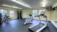 This fitness center is a dedicated, single-floor exercise facility designed to support a well-rounded workout routine. The space is outfitted with a selection of True-brand cardio equipment, including a treadmill, an elliptical trainer, and a stationary cycling bike, all positioned to take advantage of the natural light streaming in through multiple windows along the walls. A multi-station cable weight machine is situated in the corner, providing users with a range of strength training options. The flooring consists of interlocking rubber mat tiles, offering a durable, cushioned surface well-suited for exercise and equipment placement. Overhead fluorescent lighting ensures the room remains well-lit throughout the day. The walls are painted in a neutral beige tone, contributing to a clean and functional aesthetic. The windows, framed in light wood trim, offer views of surrounding greenery, adding a refreshing connection to the outdoors. The overall layout is practical and open, allowing users to move freely between equipment. This facility provides a convenient and comprehensive environment for cardiovascular fitness and strength training within a compact, efficiently organized space.
