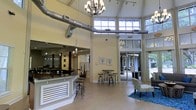 Located at 8800 Pine Forest Rd in Pensacola, Florida, this single-story space makes a striking first impression with its dramatic vaulted ceilings that soar overhead, creating an expansive and airy atmosphere. Exposed industrial-style ductwork runs prominently through the upper reaches of the main area, adding a modern, urban design element that contrasts beautifully with the warm, neutral tones of the walls and light hardwood flooring throughout.

The main gathering area is anchored by an elegant chandelier and features a reception or service counter with a sleek granite top and decorative paneling, accompanied by industrial-style bar stools. High clerestory windows line the upper walls, flooding the interior with generous natural light and offering views of the lush, tree-lined surroundings outside.

A comfortable seating arrangement with blue accent rugs defines a welcoming lounge zone, while bistro-height tables and stools provide casual gathering spots. Large glass sliding doors open toward an outdoor area, seamlessly blending interior and exterior spaces. An adjacent wing offers a more intimate setting with additional seating and décor, complementing the open grandeur of the main hall. The overall ambiance is bright, modern, and inviting, well-suited for community gatherings or professional use.
