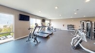 Located at 96 Coco Plum Dr in Marathon, Florida, this single-floor fitness center offers a comprehensive exercise facility designed to accommodate a wide range of workout routines. The space is laid out as one open exercise room with durable rubber flooring throughout, providing both safety and functionality for various types of training.

The cardio zone is well-stocked with multiple treadmills, a stationary spin bike, an elliptical, and a rowing machine, arranged to maximize floor space while maintaining comfortable clearance between equipment. A dedicated strength training area features a multi-station cable machine, additional selectorized weight machines, and a dumbbell rack with a full range of free weights. A stability ball, medicine balls, and a workout bench round out the functional training options.

Large windows along the perimeter allow ample natural light to fill the space and provide views of the surrounding Marathon landscape. Recessed ceiling lighting supplements illumination throughout. Multiple wall-mounted flat-screen televisions are positioned throughout the room for user convenience. A bold orange accent wall adds visual energy to the space. An adjacent locker area with built-in cubbies and seating provides a functional transition zone near the entrance, which features glass-panel doors leading to the exterior.