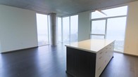 This high-rise unit is defined by its striking open-concept layout and an abundance of natural light. Floor-to-ceiling corner windows wrap around the main living area, offering expansive, panoramic views of the surrounding cityscape and waterfront — a standout feature that immediately sets the tone for the space. The exposed concrete ceiling and cylindrical concrete column lend an industrial-modern aesthetic, complementing the clean, minimalist design throughout.

Dark hardwood flooring runs across the main living area, grounding the space with warmth and contrast against the bright, airy atmosphere created by the oversized windows. At the center of the space sits a substantial kitchen island with a crisp white countertop and dark wood cabinetry, providing generous storage through multiple drawers and serving as a natural focal point for both cooking and entertaining.

The overall ambiance is sleek and contemporary, with a neutral palette that allows the dramatic city views to take center stage. The open floor plan provides a flexible, unobstructed layout suitable for a variety of configurations. The combination of raw architectural elements, modern finishes, and breathtaking elevated views makes this a visually compelling and functionally versatile space.