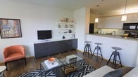 This well-designed single-floor residence offers a thoughtfully arranged open-concept layout that connects the living and kitchen areas into one cohesive, functional space. The living area is anchored by rich hardwood flooring, a sleek dark media console, and floating white shelves that provide both display space and a clean, modern aesthetic. A bold geometric-patterned area rug adds visual interest, complementing the contemporary glass coffee table and comfortable seating arrangement. Decorative accents in warm tones bring personality to the neutral, light-filled walls. The kitchen is seamlessly integrated into the open floor plan, featuring dark countertops with a stone finish, white cabinetry, and a peninsula breakfast bar outfitted with industrial-style adjustable bar stools. A pendant light fixture above the bar adds a warm, focused glow that enhances the space's modern character. The kitchen is equipped with full-sized appliances, and the cabinetry provides ample storage. Throughout the space, the combination of clean lines, neutral tones, and carefully chosen furnishings and finishes creates a cohesive and inviting atmosphere that is both practical and visually appealing.