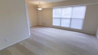 This single-floor space offers a clean and functional layout designed for comfortable everyday living. The main living area is generously sized, featuring wall-to-wall plush carpeting in a warm neutral tone that adds softness underfoot and contributes to the overall calm, understated aesthetic. Cream-colored walls provide a versatile backdrop, allowing the space to feel open and adaptable to a variety of furnishing styles. A large multi-pane window fitted with horizontal blinds floods the room with natural light, creating a bright and airy atmosphere throughout the day. A pendant light fixture suspended from the ceiling adds a subtle decorative touch while providing centralized overhead illumination. A floor-level vent indicates a dedicated heating and cooling system, ensuring year-round comfort. A doorway visible along the wall suggests additional rooms or access points, hinting at a connected and practical floor plan. The overall ambiance of the space is neutral, clean, and move-in ready, offering a welcoming blank canvas well-suited for a range of uses and interior design preferences.