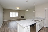 This single-story residence features a thoughtfully designed open-concept layout, connecting the living area and kitchen in a cohesive, functional space. The living area is finished with durable wood-look plank flooring in a warm, neutral tone, complemented by soft taupe walls and a flush-mount ceiling light fixture. Large windows and a glass exterior door allow natural light to fill the space, creating a bright and welcoming atmosphere.

The kitchen is a standout feature, centered around a wide granite-topped island with pendant lighting overhead. White cabinetry offers ample storage, while stainless steel appliances—including a refrigerator with water dispenser, range, and microwave—provide a modern, polished finish. Recessed lighting adds further illumination throughout the kitchen area.

The bedroom is finished with plush carpeting, neutral-toned walls, a ceiling fan with integrated light, and a window overlooking the exterior, ensuring comfortable ventilation and natural light.

The bathroom features large-format tile flooring, a walk-in shower with chrome fixtures, a white toilet, and a vanity with granite countertops and white cabinetry. A dedicated laundry closet houses a full-size top-load washer and dryer, adding everyday convenience to the unit.