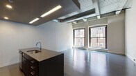 This single-floor loft-style unit showcases an industrial-modern aesthetic with exposed ductwork running across the ceiling, wide-plank hardwood flooring, and large arched windows that frame views of the surrounding urban streetscape and flood the open living area with natural light. The expansive open-plan living and kitchen area forms the heart of the unit, anchored by a substantial dark-finished kitchen island with an integrated sink and stone countertop. The kitchen is fully equipped with stainless steel appliances, including a range, microwave, and refrigerator, complemented by floor-to-ceiling dark cabinetry and a clean white tile backsplash. Recessed and linear flush-mounted lighting fixtures provide consistent illumination throughout. A separate, carpeted bedroom offers a quieter retreat, featuring exposed ductwork at the ceiling that maintains the loft's industrial character. The bathroom is finished with large-format dark tile flooring, a dark-cabineted vanity with a stone countertop, a wall-mounted mirror with sconce lighting, and a full soaking tub with a shower. A frosted sliding door adds a contemporary touch to the bathroom entry. Overall, the unit combines raw industrial elements with refined modern finishes to create a functional and visually striking living space.