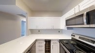 This single-story space includes a well-appointed kitchen that serves as a functional and stylish centerpiece. The kitchen features crisp white shaker-style cabinetry extending to the upper walls, providing ample storage. Light grey subway tile backsplash lines the walls between the upper and lower cabinets, adding a clean, contemporary touch. The countertops are white quartz, offering a bright, durable workspace that wraps around in an L-shaped configuration. Appliances include a stainless steel over-the-range microwave and an electric range, complemented by a dishwasher integrated beneath the counter. A standout feature is the built-in wine cooler tucked beneath the counter, adding a refined, practical element. The neutral warm-toned walls and consistent finishes create a cohesive aesthetic throughout. A glimpse into the adjacent room reveals consistent flooring and neutral wall tones, suggesting a similarly clean and modern design language carried throughout the rest of the space.