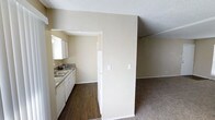 This single-floor residence offers a practical and comfortable layout designed for everyday living. The kitchen is efficiently arranged with white cabinetry providing ample storage, a stainless steel sink, granite-style countertops, and white appliances including a full-size refrigerator. The kitchen floor is finished with dark hardwood-style flooring that adds a warm, grounded contrast to the neutral cream-toned walls. A textured white ceiling runs throughout the space, adding subtle visual depth overhead. The kitchen opens toward the main living area through a half-wall partition, creating a sense of connectivity between the two spaces while maintaining a defined layout. The living area transitions to soft wall-to-wall carpeting, contributing to a cozy and inviting atmosphere. Light switch placements and standard baseboards throughout reflect a clean, well-maintained interior. The overall color palette of warm whites and beige tones creates a neutral canvas well-suited for a variety of furnishing styles. The space is straightforward in design, prioritizing functionality and ease of movement across its single level.