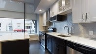 This contemporary single-floor space is defined by a cohesive modern aesthetic carried throughout its layout. A long, well-appointed corridor serves as the spine of the space, lined with crisp white walls, clean white baseboards, and rich dark hardwood flooring that runs continuously underfoot. Recessed lighting illuminates the hallway, which connects the various rooms and leads toward a bright, street-facing area at its end.

The kitchen is a standout feature, designed with a refined, functional sensibility. Dark espresso lower cabinets are paired with white upper cabinets — some with glass-front panels — creating a striking two-tone contrast. Light quartz countertops provide generous workspace, while a lustrous blue-grey mosaic tile backsplash adds texture and a subtle touch of color. Stainless steel appliances, including a range with double ovens, a mounted microwave, and a hood vent, lend a professional-grade quality to the space. A stainless undermount sink sits beneath a modern arc faucet. Large windows along the kitchen wall invite abundant natural light and offer views of the surrounding streetscape, enhancing the open and airy atmosphere. The overall ambiance is polished, modern, and thoughtfully detailed throughout.