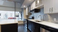 This contemporary single-floor space is defined by a cohesive modern aesthetic carried throughout its layout. A long, well-appointed corridor serves as the spine of the space, lined with crisp white walls, clean white baseboards, and rich dark hardwood flooring that runs continuously underfoot. Recessed lighting illuminates the hallway, which connects the various rooms and leads toward a bright, street-facing area at its end.

The kitchen is a standout feature, designed with a refined, functional sensibility. Dark espresso lower cabinets are paired with white upper cabinets — some with glass-front panels — creating a striking two-tone contrast. Light quartz countertops provide generous workspace, while a lustrous blue-grey mosaic tile backsplash adds texture and a subtle touch of color. Stainless steel appliances, including a range with double ovens, a mounted microwave, and a hood vent, lend a professional-grade quality to the space. A stainless undermount sink sits beneath a modern arc faucet. Large windows along the kitchen wall invite abundant natural light and offer views of the surrounding streetscape, enhancing the open and airy atmosphere. The overall ambiance is polished, modern, and thoughtfully detailed throughout.